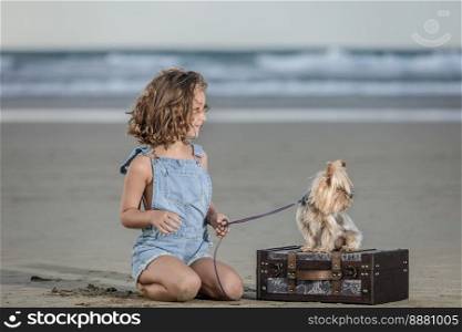 Glad girl in denim overall shorts and loyal Yorkshire Terrier dog on leash looking away while spending time on sandy Famara Beach in Lanzarote, Spain. Positive girl and dog during summer vacation on beach