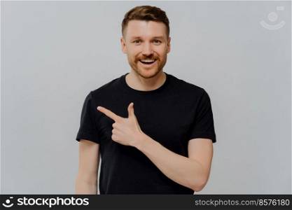 Glad handsome bearded young European man with cheerful expression shows advertisement dressed casually being in good mood suggests to use copy space advertises product on sale gives direction