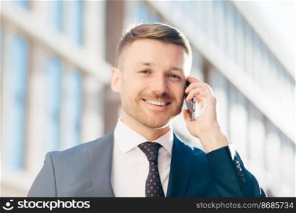 Glad handsome cheerful male entrepreneur wears formal suit, tie and white shirt, holds modern cell phone, communicates with business partner, discusses contract terms. People and occupation.