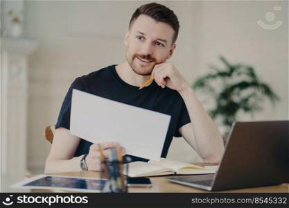 Glad handsome European businessman holds paper document poses in home interior at desktop surrounded by modern technologies checks information has remote work looks thoughtfully into distance
