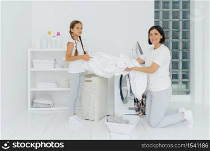 Glad housewife does washing with little adorable helper. Mother and daughter wash clothes in laundry room, load linen in washer. Woman stands on knees near washing machine. Housework concept