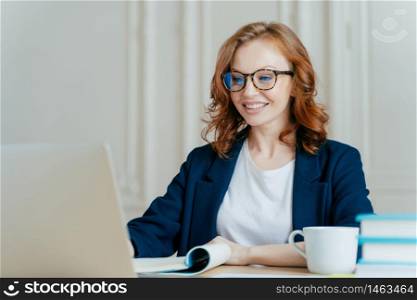 Glad lovely woman has ginger hair, positive smile, sits with laptop computer at desktop, happy to make video call or conference, communicates with investors from abroad, drinks aromatic coffee.