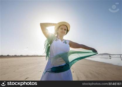 Glad middle aged female tourist with translucent scarf touching straw hat and looking at camera while dancing on sandy seashore on sunny summer day on resort. Happy woman dancing on beach