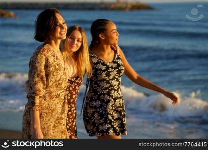 Glad multiracial female friends in sundresses strolling together on coast near waving sea while enjoying summer day in coastal area. Content multiethnic girlfriends walking on seashore