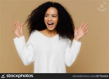 Glad overemotive woman surprised by pleasant relevation, raises hands and shows palms, focused aside, wears casual white sweater, models over brown background. Positive human emotions and feelings