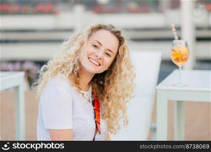 Glad pleasant looking blonde female with wavy hair, has broad shining smile, dressed in casual clothes, rests in resort place, fresh summer cocktail stands in background. People and lifestyle concept