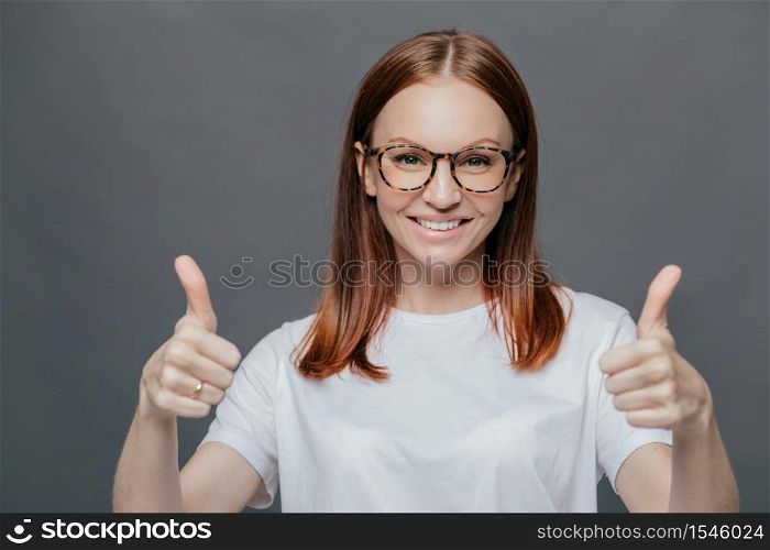 Glad positive woman with tender smile on face, has brown hair, raises two thumbs, demonstrates her approval, isolated over grey background, wears spectacles, wears casual white t shirt. Body language
