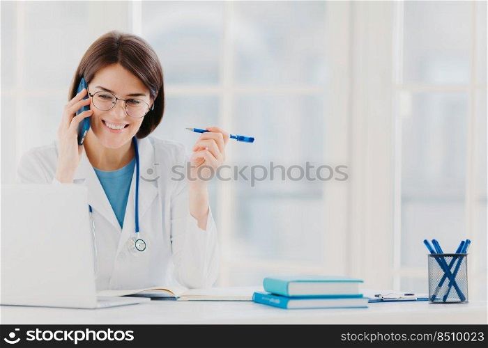 Glad professional doctor concentrated in modern laptop computer, reads useful information, has telephone conversation, discusses medical issues, sits at hospital office with notepads on table