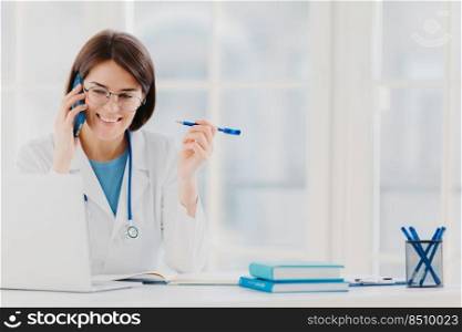 Glad professional doctor concentrated in modern laptop computer, reads useful information, has telephone conversation, discusses medical issues, sits at hospital office with notepads on table