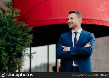 Glad prosperous businessman keeps arm folded, wears formal suit, looks aside with positive expression, rejoices success and high sales, poses outdoor against red building. Smiling smart lawyer