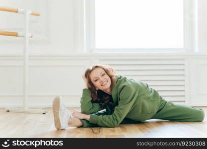 Glad red haired Caucasian young woman demonstrates good flexibility, makes leg split, prepares for ballet rehearsal, practices ballet at dance studio or fitness center, wears green sportswear.