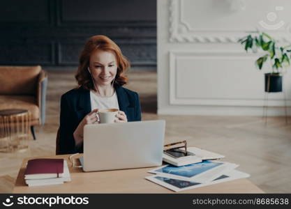 Glad red haired woman has distance job, communicates in video chat with colleagues, discusses ideas for project, enjoys online conferene uses modern laptop and accessories satisfied with friendly talk