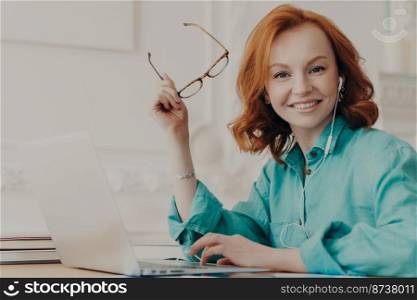 Glad red haired woman in blue shirt, works on laptop, uses earphones for watching webinar or online communication, listens courses, studies remotely from home during pandemic corona virus outbreak