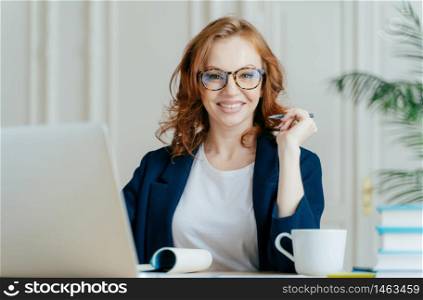Glad redhead curly woman works freelance, uses laptop computer, writes down notes in notepad, drinks coffee, wears optical glasses, being in good mood, develops business company. Work concept