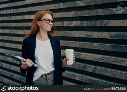 Glad redhead young woman enjoys life has successful day good mood drinks takeaway coffee holds digital tablet and notepad looks happily into distance wears formal clothes poses outdoor near wall. Glad redhead young woman drinks takeaway coffee holds digital tablet and notepad