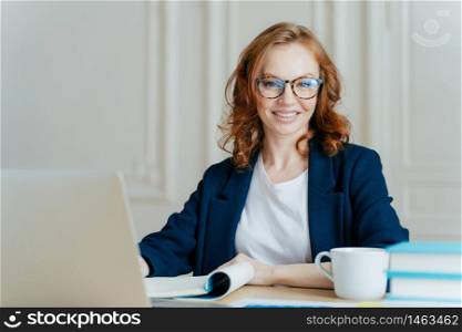 Glad smiling red haired young female concentrated on creating new business project, owns corporation, sits in front of laptop computer, wears optical glasses and elegant outfit, updates software