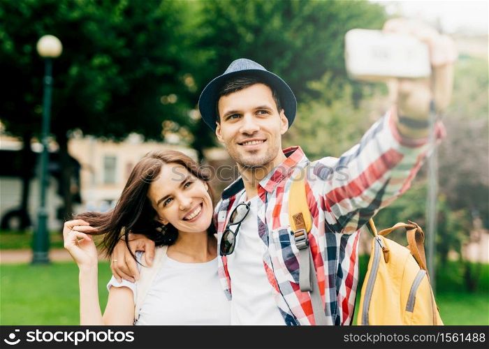 Glad tourists with rucksack, dressed casually, making selfie with their camera, posing against green nature background, having broad smiles on their faces. People, lifestyle, sightseeing concept