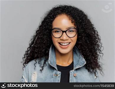 Glad woman with curly hair, wears optical glasses, denim jacket, looks straightly at camera, isolated on grey background, enjoys lovely conversation, satisfied with finished work. Facial expressions