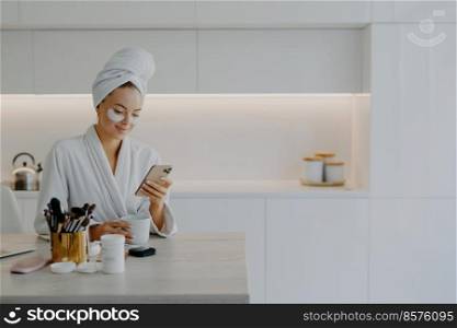 Glad young European woman dressed in bathrobe types text messages in online chat during coffee break undergoes skin care procedures applies beauty patches poses over kitchen interior at home