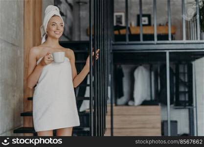 Glad young European woman smiles pleasantly, drinks coffee or tea, concentrated aside, stands wrapped in bath towel, feels refreshed after taking shower, has well cared body and healthy skin