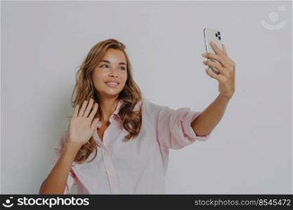 Glad young European woman waves palm shows hello gesture poses at smartphone camera smiles gladfully dressed casually has online call with friend isolated over grey background. Nice to meet you. Glad young European woman waves palm shows hello gesture poses at smartphone camera