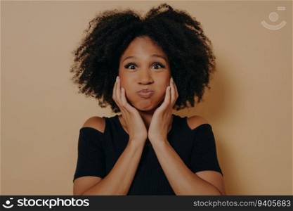 Glamorous African model with wavy hair, making a funny face by puffing out her cheeks and holding them. Indoor portrait on dark beige background with copy space.