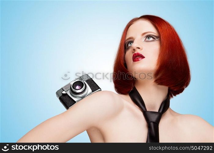 Glamorous girl, vamp style, wearing a necktie, holding an old vintage photo camera on her pretty shoulder on blue background.