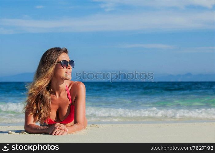 Glamorous long haired young woman in bikini and sunglasses lying on tropical beach, copy space for content. Woman lying on tropical beach
