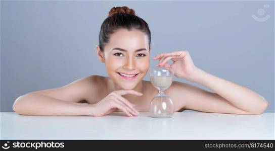 Glamorous model holding hourglass in beauty concept of anti-aging skincare treatment for woman. Young asian girl portrait with perfect smooth clean skin and flawless soft makeup in isolated background. Glamorous beautiful woman with hourglass as anti-aging skincare concept