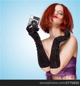 Glamorous posing girl, wearing long gloves, holding an old vintage photo camera and grimacing on blue background.