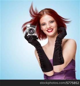 Glamorous smiling girl, wearing long gloves, holding an old vintage photo camera, and saying cheese on blue background.