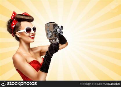 Glamorous vintage pin-up girl filming movie with an old retro cinema 8 mm camera, standing on colorful abstract cartoon style background.