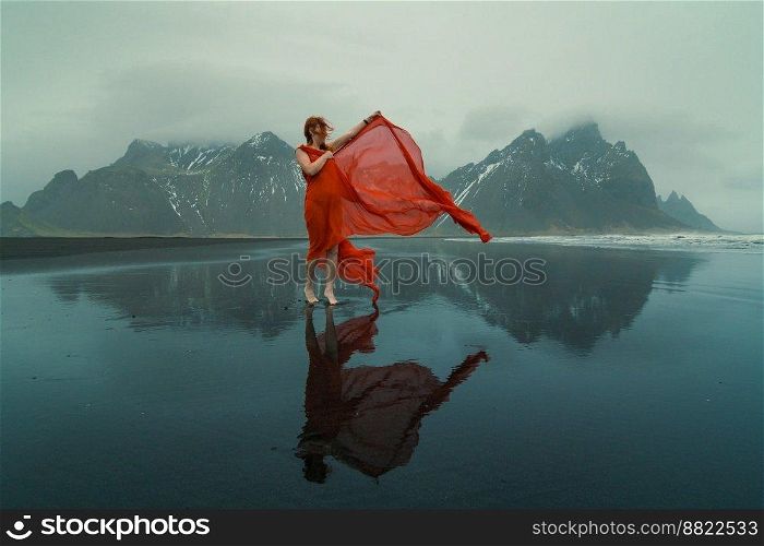 Glamorous woman in red dress on northern beach scenic photography. Picture of person with mountains on background. High quality wallpaper. Photo concept for ads, travel blog, magazine, article. Glamorous woman in red dress on northern beach scenic photography