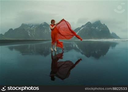 Glamorous woman in red dress on northern beach scenic photography. Picture of person with mountains on background. High quality wallpaper. Photo concept for ads, travel blog, magazine, article. Glamorous woman in red dress on northern beach scenic photography