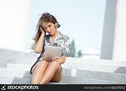 Glamorous young woman with vintage music headphones and a take away coffee cup, surfing internet on tablet pc, listening to the music and sitting on stairs against urban city background.
