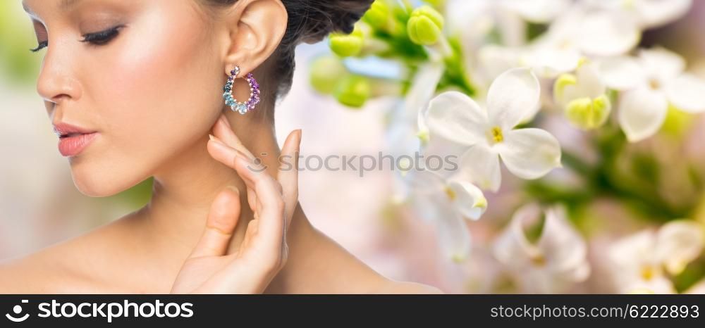 glamour, beauty, jewelry and luxury concept - close up of beautiful woman face with earring over natural spring lilac blossom background