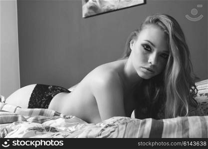 glamour close-up portrait of stunning woman with freckles and long blonde hair lying on bed with only lace black panties and nude body. She is looking in camera . monochrome image