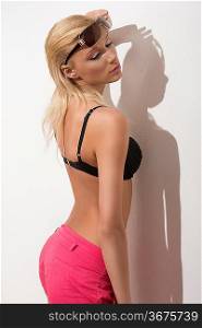 glamour summer portrait of sexy blonde girl with wet blonde hair, bra, shorts and sunglasses. She showing her back near white wall