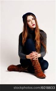 Glamourous portrait of the young beautiful woman in leather boots and autumn clothes posing over white background, sitting on the floor. Studio shot. Hipster style.