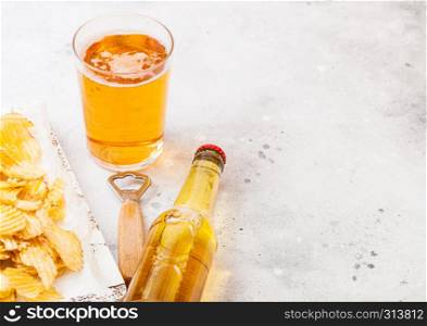 Glass and bottle of craft lager beer with potato crisps snack in vintage wooden box and opener on stone kitchen background. Beer and snack.