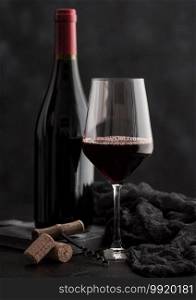 Glass and bottle of red wine with corks and vintage corkscrew with dark cloth on black background.