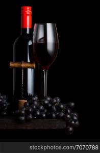 Glass and bottle of red wine with dark grapes and vintage corkscrew opener and cork on wooden board on black