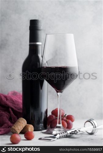 Glass and bottle of red wine with red cloth and corks with steel corkscrew and grapes on light stone background.