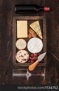 Glass and bottle of red wine with selection of various cheese in wooden box and grapes on wooden background. Blue Stilton, Red Leicester and Brie Cheese and knife.