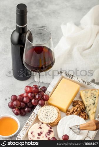 Glass and bottle of red wine with selection of various cheese in wooden box and grapes on light background. Blue Stilton, Red Leicester and Brie Cheese with Cheddar and knife.