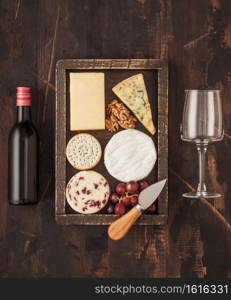 Glass and bottle of red wine with selection of various cheese in wooden box and grapes on wooden background. Blue Stilton, Red Leicester and Brie Cheese and knife.