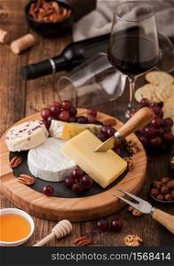 Glass and bottle of red wine with selection of various cheese on the board and grapes on wooden table background. Blue Stilton, Red Leicester and Brie Cheese and knife with linen kitchen cloth.
