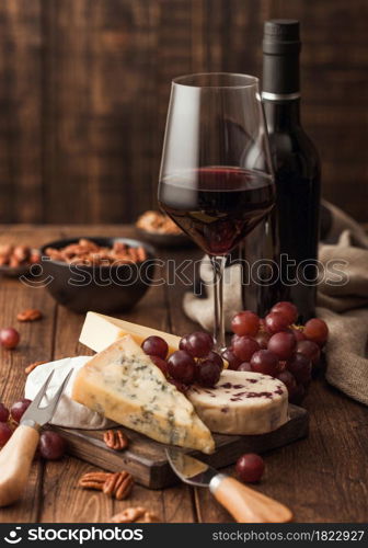 Glass and bottle of red wine with selection of various cheese on the board and grapes on wooden table background. Blue Stilton, Red Leicester and Brie Cheese and knife.