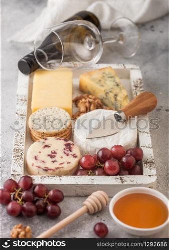 Glass and bottle of white wine with selection of various cheese in wooden box and grapes on wooden background. Blue Stilton, Red Leicester and Brie Cheese and Cheddar with knife.
