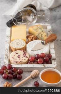 Glass and bottle of white wine with selection of various cheese in wooden box and grapes on wooden background. Blue Stilton, Red Leicester and Brie Cheese and Cheddar with knife.